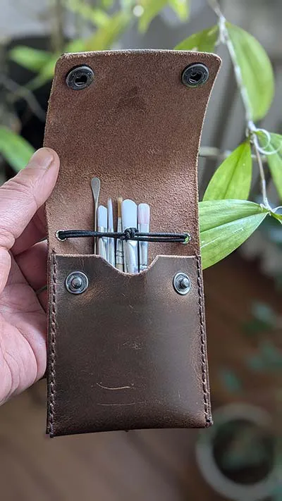 Slim brown leather pouch with tools inside, held open in front of a plant